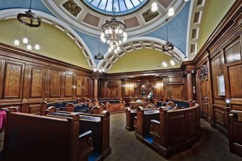 Victorian council chamber interior with ornate wooden panelling, leather benches, a domed skylight and chandelier